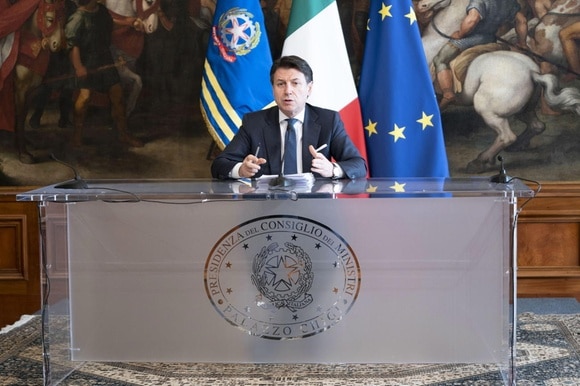 Uomo in abiti formali seduto a un pulpito con scritta Presidenza consiglio dei ministri Bandiera italiane ed europea dietro