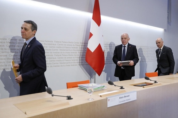 Tre uomini in abiti formali si apprestano a sedersi a un tavolo di conferenza stampa; bandiera svizzera sul fondo