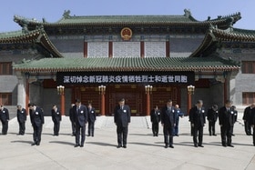 Gruppo di uomini in abito formale e scuro disposti in ranghi fanno un inchino; dietro, costruzione cinese