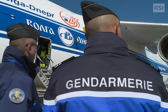 Due gendarmi francesi di spalle sorvegliano personale che cariga un velivolo cargo con scritte anche in cirillico