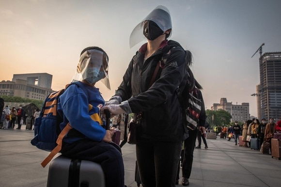 Una mamma e un bambino bardati con visiera mascherina e altri strumenti protettivi su un piazzale con gente in coda