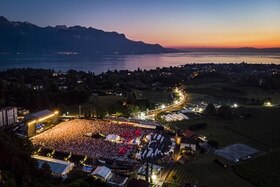 Vista aerea di un concerto al crepuscolo, in un parco nei pressi di un lago; si distinguono palco e pubblico