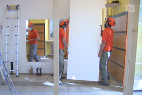 Tre uomini in abiti da lavoro, con maglietta arancione, montano pannelli di separazione in una stanza.