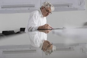 Un uomo con capelli bianchi e occhiali traccia qualcosa a matita su un ampio tavolo da lavoro; disegni tecnici alle pareti