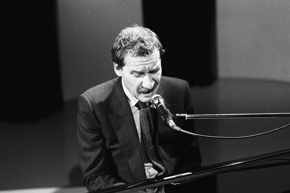 Un uomo in giacca e cravatta seduto al pianoforte, suona e canta in un teatro o studio televisivo.
