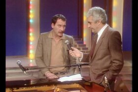 Due uomini, di cui uno in cravatta e che regge un microfono, parlano in piedi accanto a un pianoforte
