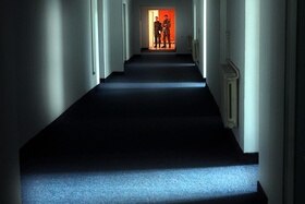 Lungo corridoio illuminato solo da fasci di luce filtranti da porte aperte; in fondo, due militari