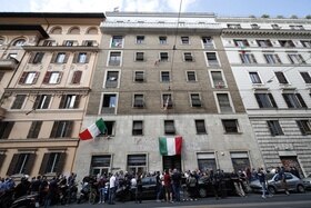 Gruppo di persone lungo il marciapiede davanti a stabile di 6 piani di inizio 900 con bandiere italiane esposte