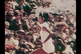 Un atleta in tenuta bianca corre in salita reggendo una fiaccola; dietro, pubblico da stadio