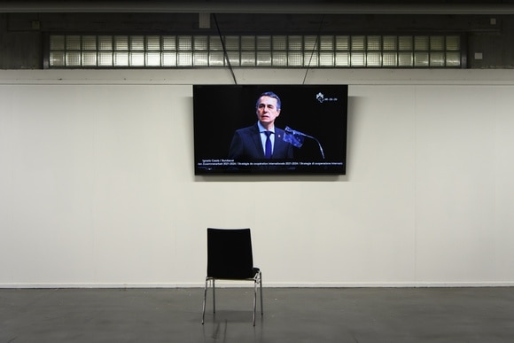 Un uomo in abito formale, visto su uno schermo sulla parete vuota di una grande sala, parla a un microfono
