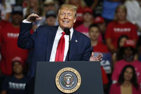 Trump al pulpito con effigie della presidenza USA, in abito blu e cravatta rossa, fa un gesto stile limitare ; pubblico dietro
