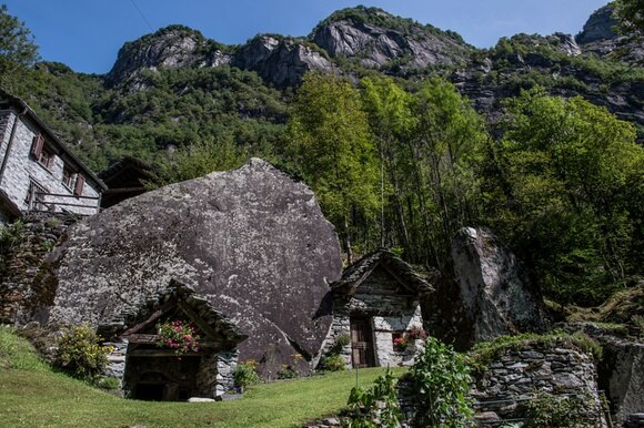 Casa in pietra costruita sotto un macigno ai piedi di un ripido pendio, con bosco e vetta rocciosa