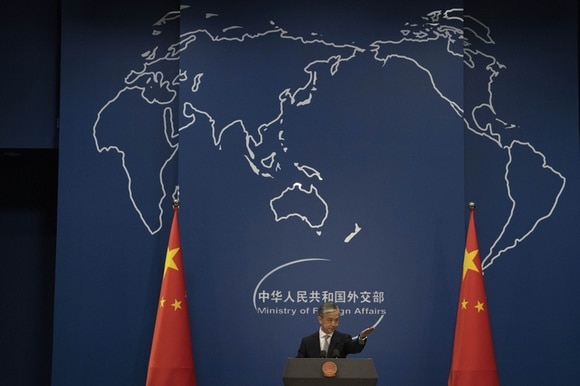 Uomo in abiti formali parla da un pulpito; dietro di lui bandiere cinesi e immagini del globo con Cina al centro