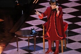 Dona anziana in abito rosso e stivali alti su palcoscenico illuminato a rombi, parla a un vecchio telefono seduta a un tavolino