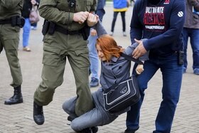 Piazza con manifestazione; donna strattonata da uomo in tuta mimetica e altro uomo in jeans e felpa
