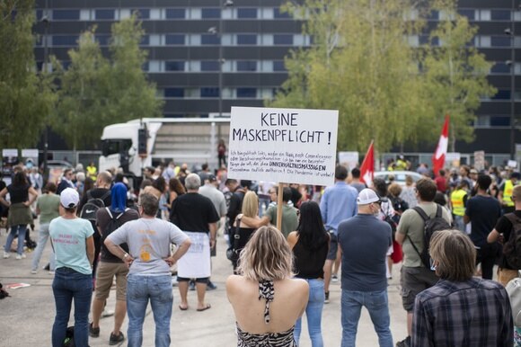 Persone in una ampia piazza viste di schiena durante una manifestazione, una ragazza regge un cartello No mascherine