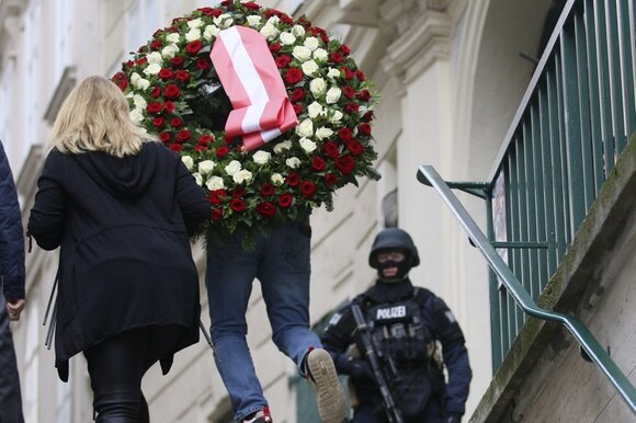 due persone portano una corona mortuaria sotto lo sguardo di un poliziotto