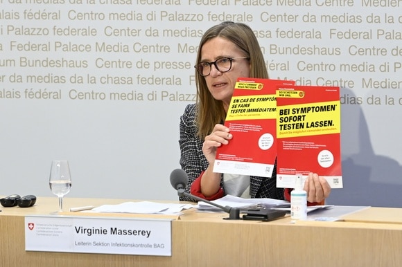Virginie Masserey al tavolo di conferenza stampa mostra due manifesti che raccomandano i test