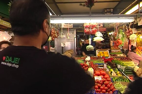 Banco di mercato con frutta e verdura. Di spalle, un giovane con maglietta con scritto www.daje.shop