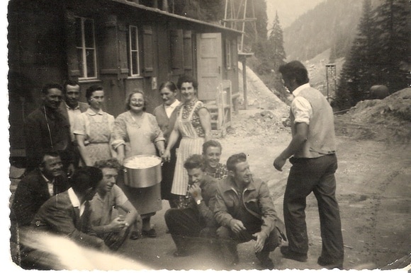 b/n Tre giovani donne e un anziana posano con un pentolone accanto a un gruppo di operai fuori da una baracca