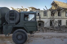 Camion militare transita davanti a fila di case contigue parzialmente distrutte