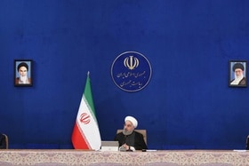 Rouhani seduto a un tavolo con accanto bandiera iraniana; fondo blu e immagini incorniciate di Khomeini e Khamenei
