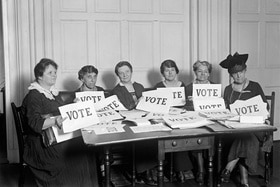 donne sedute a un tavolo con cartelloni con la scritta voto