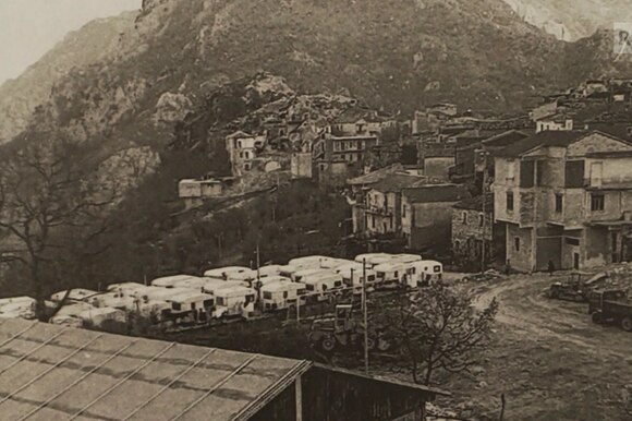 Immagine di un villaggio ai piedi di una montagna con, ai margini, una fila di roulotte parcheggiate.