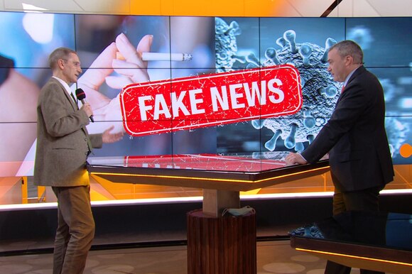 Due uomini si parlano in studio televisivo; alle loro spalle immagine illustrativa con scritta Fake news