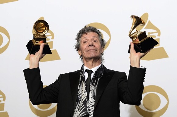 Uomo in cravatta ma casula solleva due Grammy Award mostrando un espressione soddisfatta ai fotografi
