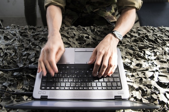 Militare lavora a un computer portatile appoggiato su telo mimetico.