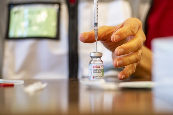 Mano di infermiera (si intravvede camice) nel gesto di afferrare una fiala di vaccino Moderna per estrarne una dose