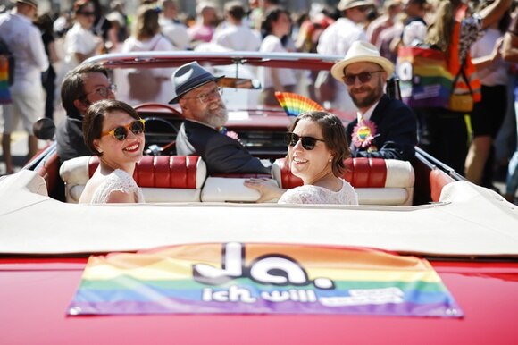 Su un auto decapottabile una coppia lesbica con la bandiera arcobaleno con la scritta, Si, lo voglio.