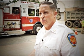 uomo con capelli grici e camicia bianca con stemma dei pompieri sulla spalla sinistra. dietro di lui un camion dei pompieri