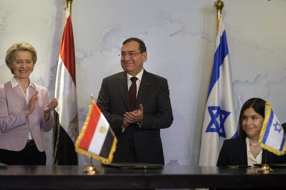 un uomo e una donna in piedi sorridono, un altra donna è seduta. davanti e dietro a loro le bandiere egiziana e israeliana