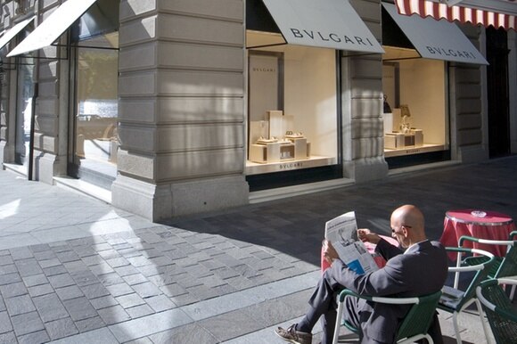 uomo seduto al tavolino esterno di un bar legge un giornale di fronte a un negozio di lusso