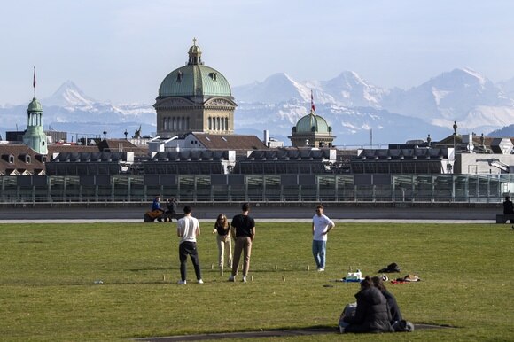 Giovani in pimo piano che giocano su un prato e dietro le cupole di Palazzo federale e le Alpi bernesi.